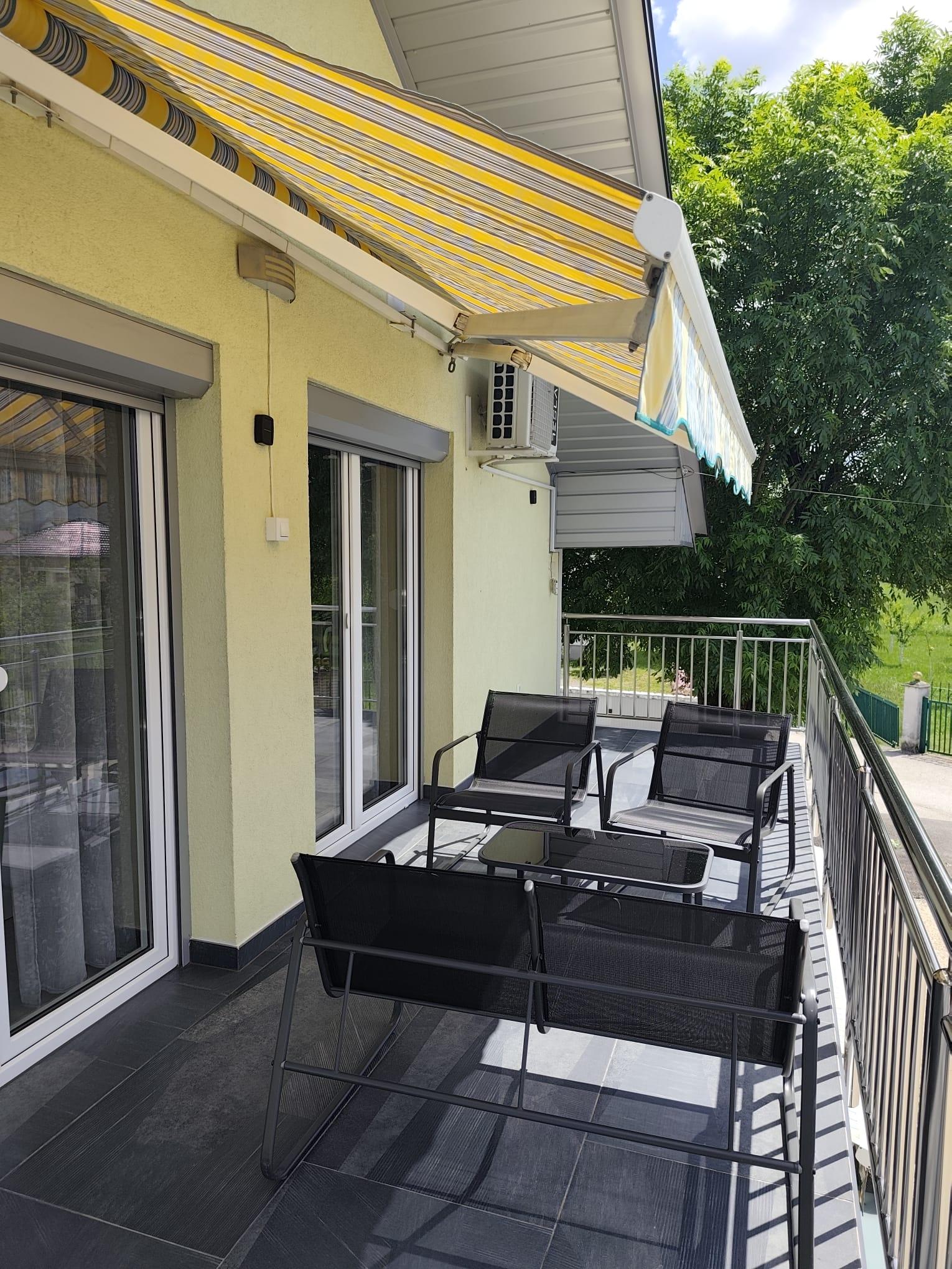 Dugi balkon sa sivim pločicama, crnim mrežastim sjedištima, staklenim stolom, prugastom izvlačnom tendom, metalnom ogradom i drvećem u pozadini.