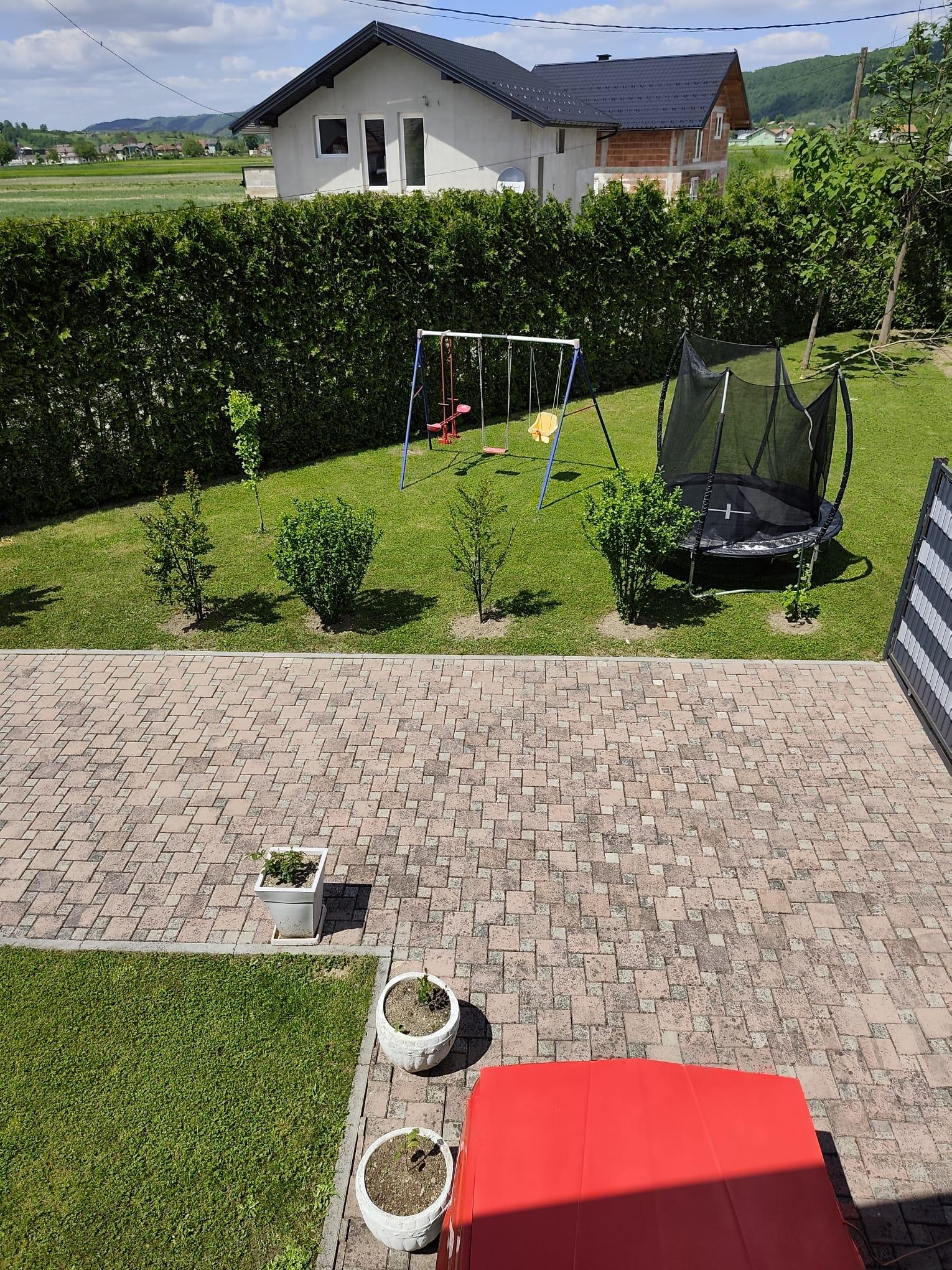 Travnjak okružen živicom s okvirom ljuljačke s tri sjedala, okruglim trampolinom s mrežom, mladim drvećem i grmljem, popločanom površinom, poljima i šumovitim brdima u daljini.