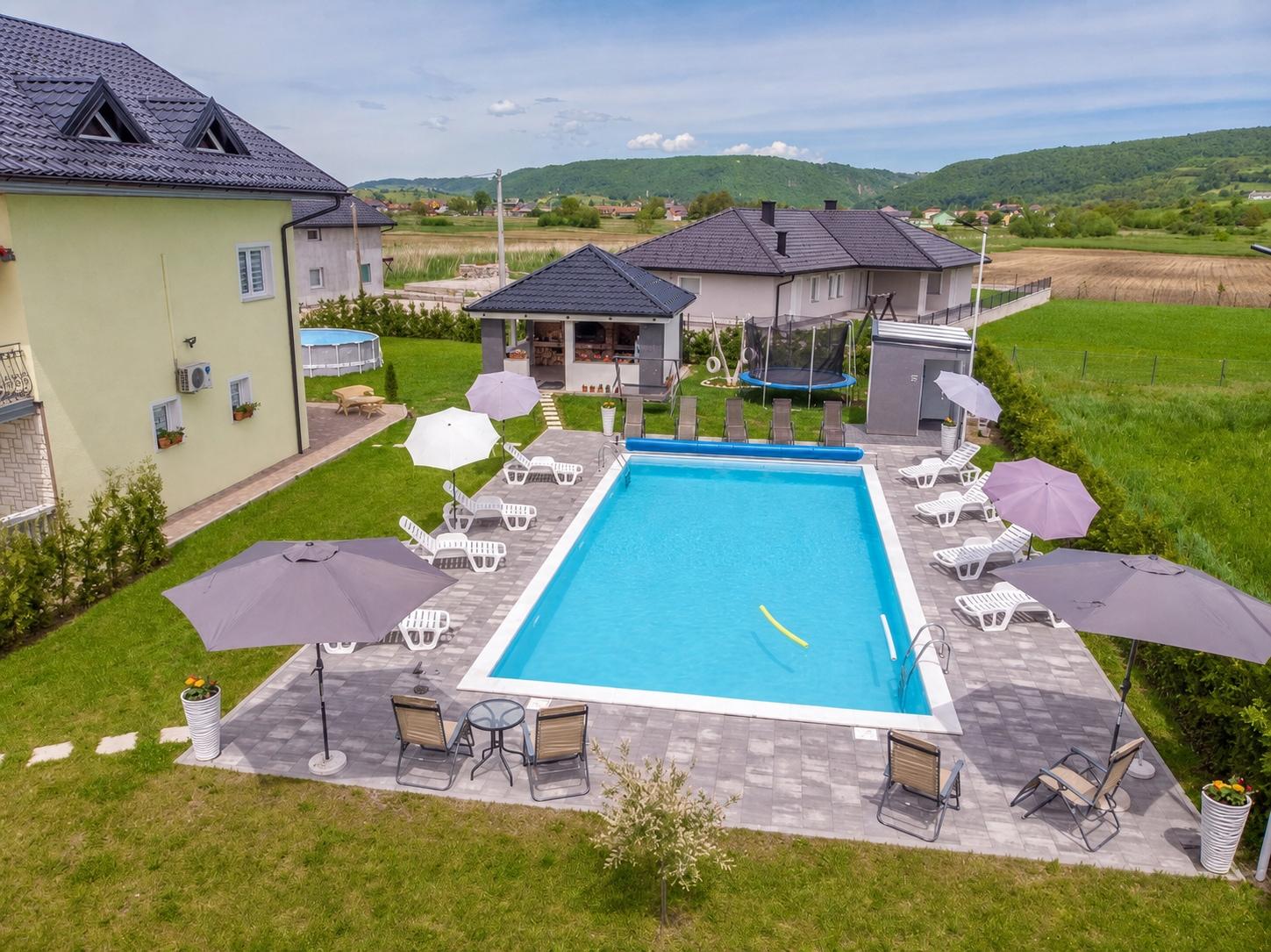 Pogled iz zraka na vilu, bazen, travnjak, ležaljke, natkriveni paviljon i okolni krajolik.
