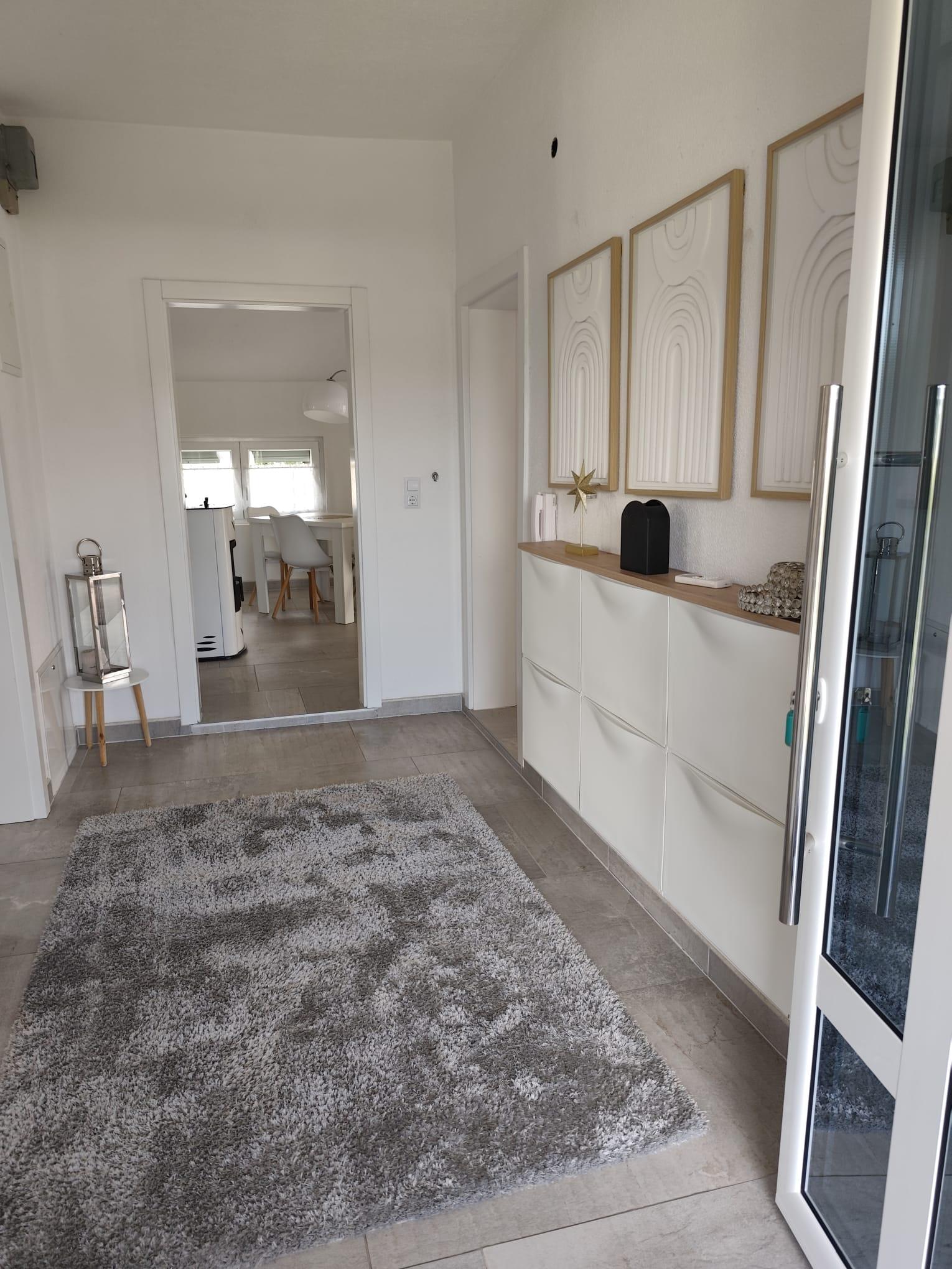 Heller weißer Eingangsbereich mit grauem Fliesenboden, gemustertem Teppich, weißem Sideboard und Durchgang zum Essbereich.