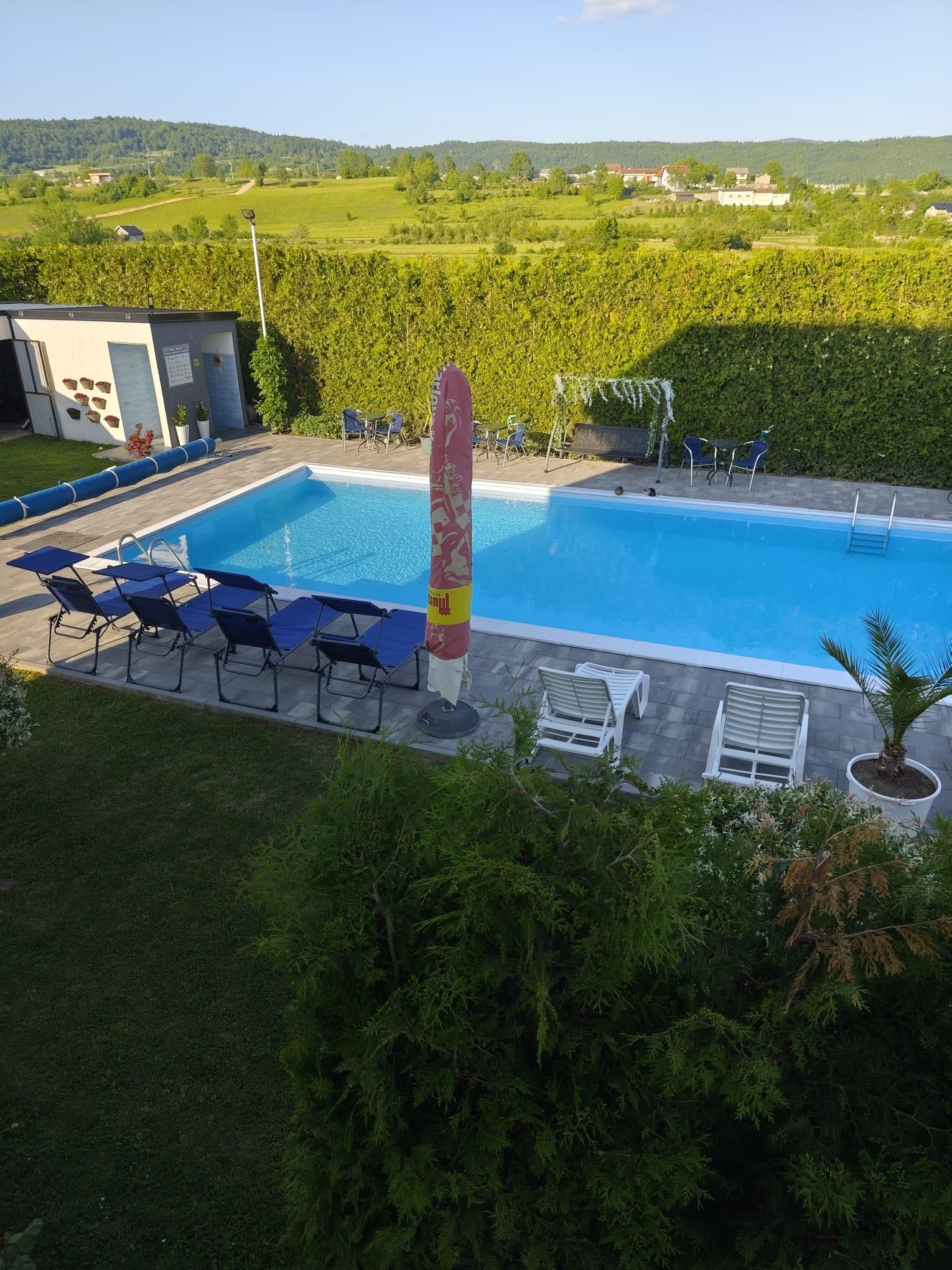 Weiter erhöhter Blick auf den Pool, die Liegestühle und die grüne Landschaft dahinter.